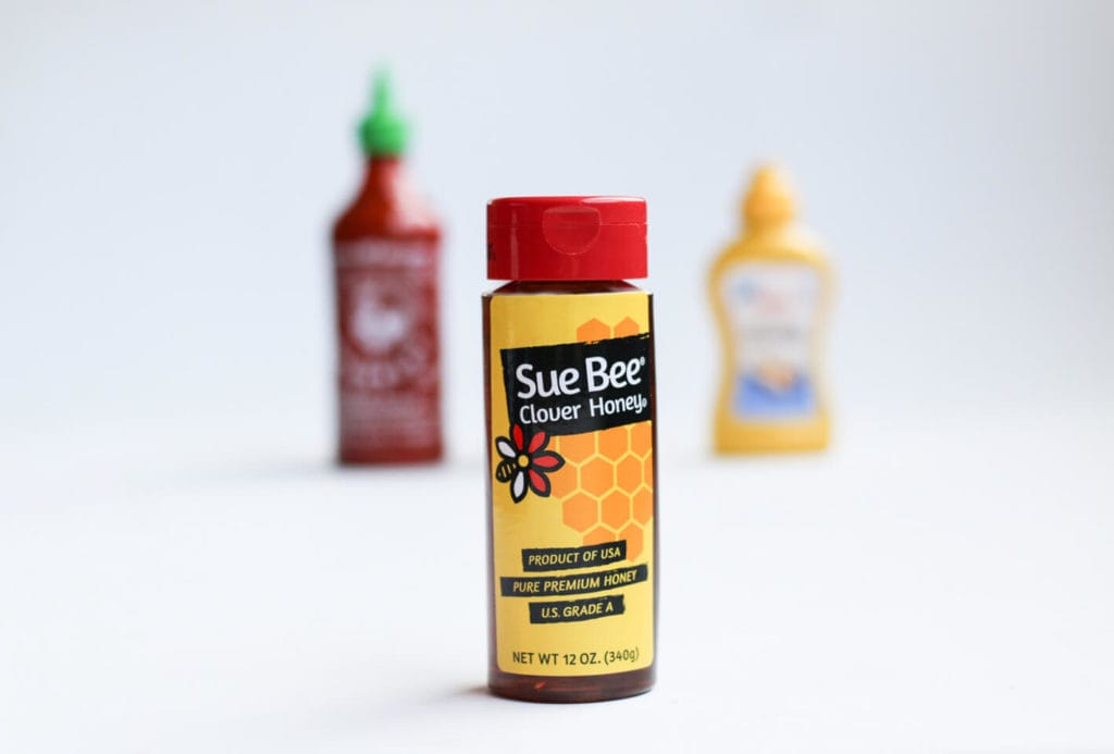 Picture of a bottle of Sue Bee Honey with Sriracha and mustard in the background.
